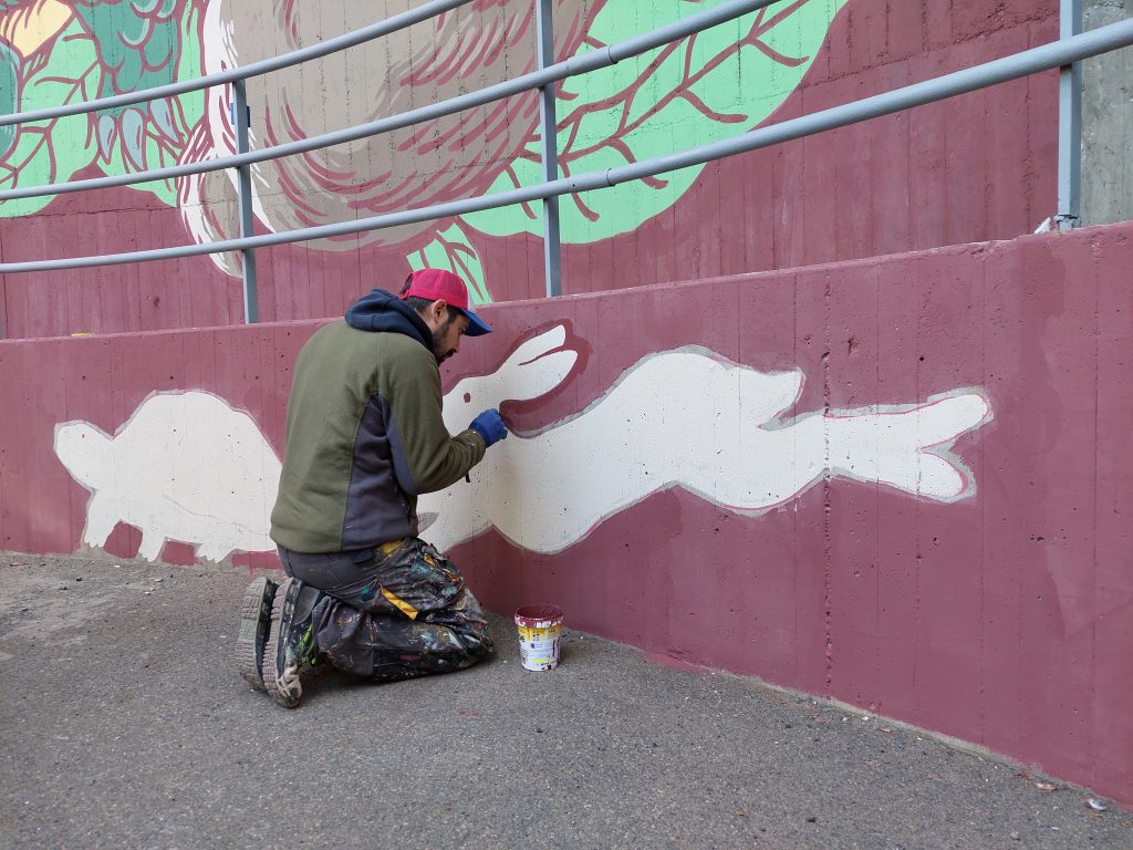 Lucamaleonte dipinge la Tartaruga e la lepre sul  muro semicircolare di una grande caldaia nel quartiere Vigne Nuove in via Dina Galli 8 